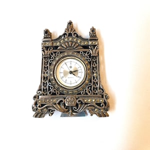 Pode incluir: Um relógio de tom prateado, adornado com um movimento de quartzo. O relógio tem a forma de uma pequena casa com um design decorativo e um mostrador branco.