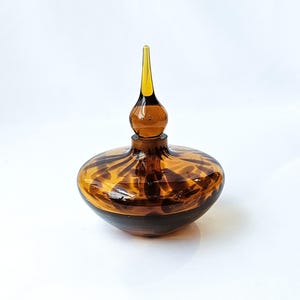 May include: A decorative glass perfume bottle with a brown and amber color scheme. The bottle has a rounded base and a long, pointed stopper. The glass has a speckled pattern, and the stopper is a lighter amber color.
