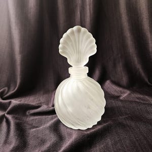 May include: A frosted glass perfume bottle with a seashell-shaped stopper. The bottle has a round, swirled design. The background is a dark purple fabric.