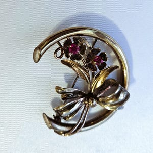 May include: A gold and silver tone brooch with a floral design and two red gemstones. The brooch is shaped like a horseshoe.