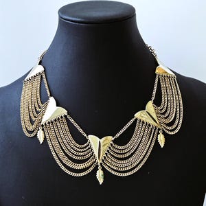 May include: A gold-colored statement necklace with a layered chain design. The necklace features geometric shapes and cascading chains, with small leaf-shaped pendants. The jewelry is displayed on a black mannequin.