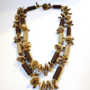Peut inclure: Un collier à double rang de perles brunes et blanches avec des accents dorés. Le collier est fait de bois, de métal et de coquillages.
