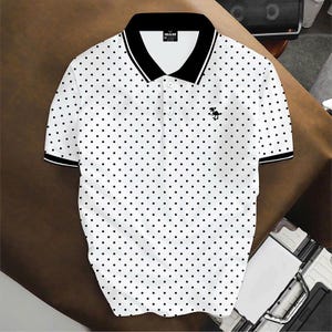 May include: A white short-sleeved polo shirt featuring a black polka dot pattern throughout. The shirt has a solid black collar with white trim, a three-button placket, and a small black dinosaur graphic embroidered on the left chest area.