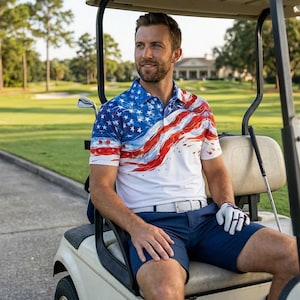 以下が含まれることがあります： 赤と青のアクセントが施された、愛国的なアメリカ国旗のデザインの白いポロシャツ。シャツはネイビーブルーのショーツと白いベルトと組み合わせています。その人はゴルフグローブを着用し、ゴルフカートに座っています。