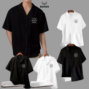 Könnte beinhalten: Schwarze und weiße kurzärmelige Button-Down-Hemden mit dem Text "YOUR DESIGN HERE" auf der Brust. Die Hemden werden auf Kleiderbügeln präsentiert. Das Markenlogo "BEAVER" befindet sich oben im Bild.
