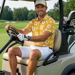 May include: A yellow polo shirt with a white fried egg pattern, white shorts, a white and yellow baseball cap with the letters OG, a white golf glove, and grey and white trainers. The attire is worn while seated in a golf cart on a course.