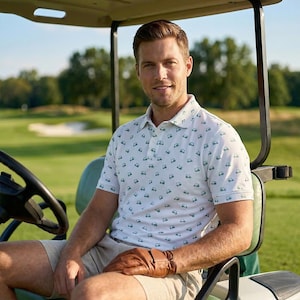 May include: A man wearing a white polo shirt with a small green golf cart pattern, khaki shorts, and brown leather gloves, sitting in a golf cart. The background shows a green golf course and trees.