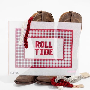 May include: A needlepoint canvas with the words "ROLL TIDE" in red letters, set against a red and white gingham pattern. Brown cowboy boots are positioned behind the canvas. Red, white, and gray yarn are in the foreground.
