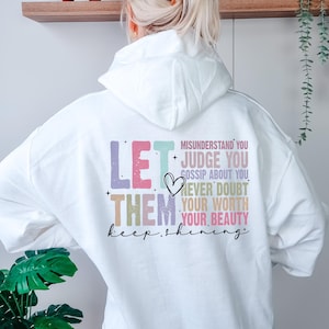 Könnte beinhalten: Weißer Kapuzenpullover mit dem Text "LET THEM keep shining" in Pastellfarben. Auf der Rückseite des Hoodies stehen außerdem die Sätze "MISUNDERSTAND YOU, JUDGE YOU, GOSSIP ABOUT YOU, NEVER DOUBT YOUR WORTH, YOUR BEAUTY."
