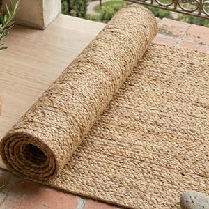 May include: A rolled-up, natural fibre rug with a woven texture lies on a brick patio. The rug is a light brown colour. Potted plants and woven slippers are also visible. The scene suggests an outdoor living space.
