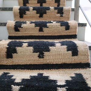 Op de afbeelding: Trapmatten met een geometrisch zwart patroon op een natuurlijke beige achtergrond. De matten zijn gemaakt van geweven materiaal en zijn geïnstalleerd op betonnen trappen. Het ontwerp heeft herhalende vormen en lijnen.