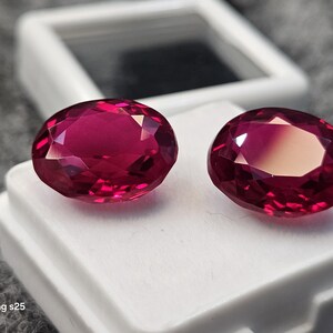 Peut inclure: Deux pierres précieuses ovales d'un rouge magenta profond avec des surfaces facettées qui reflètent la lumière. Les pierres sont placées côte à côte sur une boîte de présentation rectangulaire blanche avec un couvercle transparent.