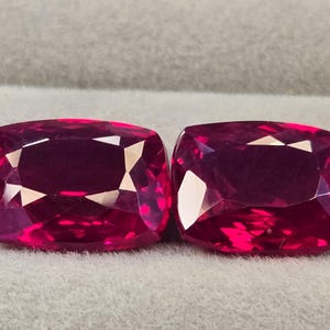 Può includere: Due pietre preziose taglio cuscino di un colore rosso intenso e vibrante. Ogni pietra presenta una superficie sfaccettata che riflette la luce, creando riflessi luminosi sulla parte superiore. Le gemme sono posizionate fianco a fianco, trasparenti.