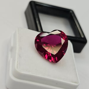 May include: A heart-shaped, faceted gemstone in shades of deep red and pink rests on a white surface. The gemstone is displayed in a white box, with a black display case in the background. The gem's facets catch the light, creating a sparkling effect.