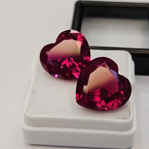 Pode incluir: Duas pedras preciosas facetadas em forma de coração de cor vermelho magenta profundo. As pedras são exibidas sobre uma base de espuma branca quadrada dentro de uma pequena caixa de joias de plástico preto com a tampa aberta.
