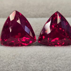 Puede incluir: Dos piedras preciosas rojas vibrantes cortadas en forma triangular con múltiples facetas. Las piedras exhiben un color carmesí profundo y saturado con un acabado de alto brillo, reflejando la luz en sus superficies geométricas con nitidez.