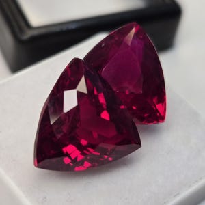 Op de afbeelding: Twee losse edelstenen in trillion-slijpvorm met een diepe, levendige magenta-rode kleur. Deze gefacetteerde stenen hebben een driehoekige vorm met scherpe punten en meerdere reflecterende oppervlakken die het licht vangen en helderheid tonen.