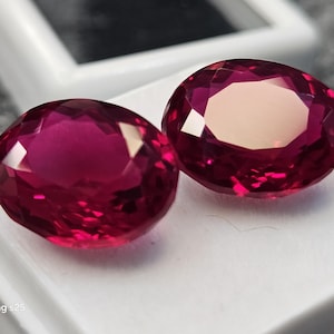 Könnte beinhalten: Zwei oval geschliffene, tief magentarote Edelsteine mit facettierten Oberflächen, die das Licht reflektieren. Die Steine liegen nebeneinander auf einem weißen Sockel. Der Text "Samsung s25" ist in der unteren linken Ecke des Bildes zu sehen.