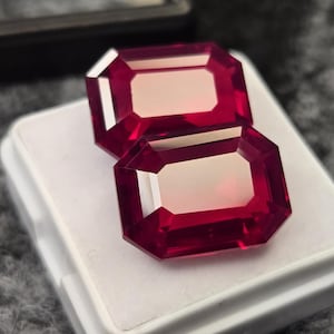 May include: Two faceted, rectangular-cut, deep red gemstones. The gems are displayed on a white surface, with a dark case visible in the background. The gems have a polished, reflective surface, showcasing their clarity and color.