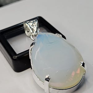 May include: A pear-shaped pendant with a large, opalescent gemstone, set in a silver-colored metal frame. The gemstone has a milky white and pale blue color. The pendant is displayed next to a black square box.