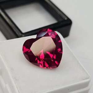 Può includere: Una gemma rossa a forma di cuore, sfaccettata, appoggiata su una superficie bianca. La gemma è esposta in una scatola bianca, con un espositore nero sullo sfondo. Il taglio crea molte superfici riflettenti.