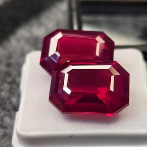 May include: Two vibrant, deep red gemstones cut in an emerald shape with faceted surfaces. The stones are positioned one slightly behind the other, displaying clear internal reflections and sharp edges, resting on a white square display base.