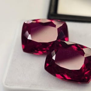 May include: Two cushion-cut, deep magenta-red gemstones with a faceted surface. The stones exhibit a high level of clarity and a reflective, polished finish, showcasing a vibrant, saturated colour profile across their symmetrical, rectangular shapes.