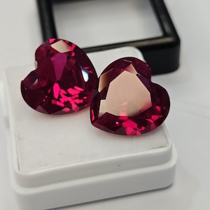 Puede incluir: Dos piedras preciosas de color rojo intenso en forma de corazón, talladas con facetas y corte brillante. Las piedras están colocadas una al lado de la otra sobre una base de exhibición blanca, mostrando sus superficies reflectantes y color vivo.