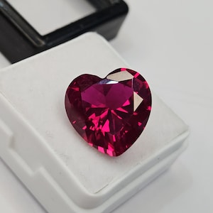 May include: A heart-shaped, deep magenta-red gemstone with a faceted surface that reflects light. The stone is displayed resting on a white foam insert inside a small, open black plastic jewelry box, showcasing its vibrant color and cut.