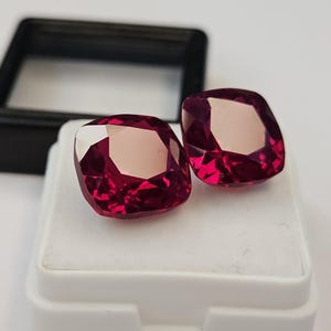 Puede incluir: Dos gemas facetadas con corte cojín y un tono rojo intenso. Las gemas se exhiben sobre una superficie blanca dentro de un recipiente de plástico blanco, con un recipiente negro en el fondo. Las gemas tienen un alto grado de claridad y brillo.