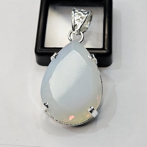May include: A teardrop-shaped pendant featuring a smooth, milky white stone with subtle iridescent highlights. The stone is secured in a silver-toned metal setting with four prongs and includes a decorative, textured silver-toned bail at the top.