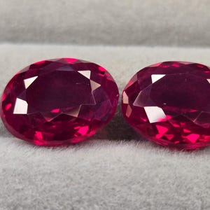 Puede incluir: Dos rubíes facetados de forma ovalada con un color rojo intenso y vibrante. Cada piedra preciosa presenta un corte preciso con múltiples facetas triangulares que reflejan la luz, creando una apariencia brillante y centelleante.