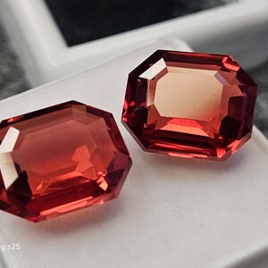 Könnte beinhalten: Zwei lose Edelsteine im Smaragdschliff mit einem tiefen, leuchtend rot-orangen Farbton. Die Steine weisen facettierte Oberflächen auf, die das Licht reflektieren und helle Glanzlichter sowie tiefe Schatten auf ihren rechteckigen, mehrfach facettierten geometrischen Formen erzeugen.