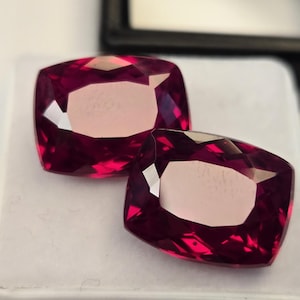 May include: Two cushion-cut, deep magenta-red gemstones with faceted surfaces. The stones exhibit a high degree of clarity and a vibrant, saturated color profile, reflecting light across their polished, angular faces while resting on a white surface.