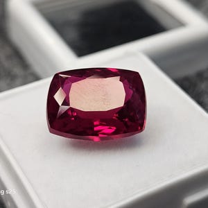 May include: A cushion-cut, faceted gemstone with a deep red hue. The gem is set on a white surface, with a blurred background. The stone's facets reflect light, creating a sparkling effect. The gem is likely a ruby or garnet.
