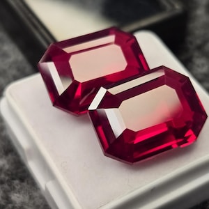 May include: Two faceted, rectangular, deep red gemstones with a lighter interior are displayed on a white surface. The gems have a brilliant cut, reflecting light. The background is a blurred, dark gray.