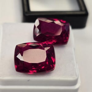 Op de afbeelding: Twee kussengeslepen, diep magentarode edelstenen met gefacetteerde oppervlakken. De stenen liggen op een witte schuimrubberen inzet in een klein zwart juwelendoosje, wat hun levendige kleur en reflecterende, geometrische patronen benadrukt.