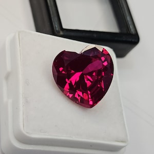 May include: A heart-shaped gemstone with a deep magenta-red colour and a faceted surface. The stone is displayed on a white square foam insert, which is part of a small black plastic display box with a clear lid visible in the background.