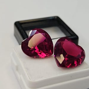 Puede incluir: Dos piedras preciosas en forma de corazón, facetadas y de color rojo magenta intenso, exhibidas sobre un inserto blanco dentro de un estuche de plástico negro. Las piedras presentan cortes geométricos precisos que crean superficies reflectantes.
