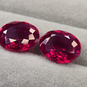 Könnte beinhalten: Zwei oval geformte, facettierte rote Edelsteine im Brillantschliff. Die Steine weisen einen tiefen, gesättigten karmesinroten Farbton und reflektierende Oberflächen auf, die zahlreiche dreieckige Facetten zeigen, welche das Licht auf ihren polierten Körpern einfangen.