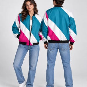 May include: A bomber jacket featuring a diagonal colour-blocked pattern in teal, white, and magenta. The jacket has a black ribbed collar, black ribbed cuffs, and a black ribbed hem, with a full-length front zip closure and long sleeves.