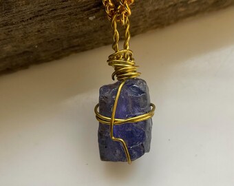 May include: A gold-colored chain necklace with a raw, dark blue-purple gemstone pendant. The gemstone is wrapped in gold wire. The necklace is displayed against a wooden background.