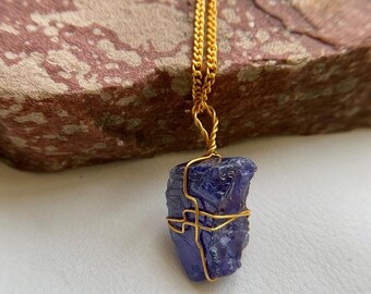 May include: A gold-colored chain necklace with a raw, dark blue tanzanite gemstone pendant. The tanzanite is wrapped in gold wire. The necklace is displayed against a textured, reddish-brown stone.