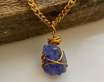 May include: A gold-colored chain necklace with a raw, blue-purple gemstone pendant. The gemstone is wrapped in gold wire, and the chain has a textured, linked design. The necklace is displayed against a light background with a wooden element.