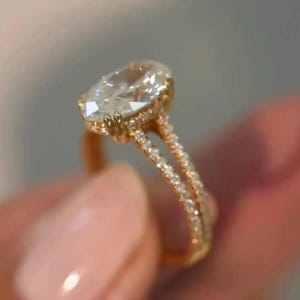 May include: A gold ring with a large, oval-cut diamond as the centerpiece. The band splits into two, each encrusted with smaller diamonds, creating a halo effect around the main stone. The ring is held up close, showcasing its intricate design.