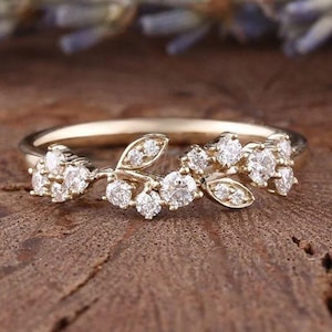 May include: A gold ring with a floral design. The ring features small, round, clear gemstones clustered to resemble flowers and leaves. The ring is set against a blurred background of lavender and a wooden surface.
