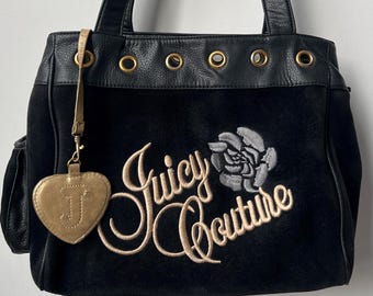 May include: Black suede tote bag with leather handles and trim. Features the embroidered text "Juicy Couture" with a rose design. Includes a gold heart-shaped charm with the letter "J".