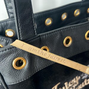 May include: Black tote bag with gold-toned grommets and handles. The bag features a gold leather tag with the inscription "I am the fairest." The brand name "Juicy Couture" is visible in gold lettering.