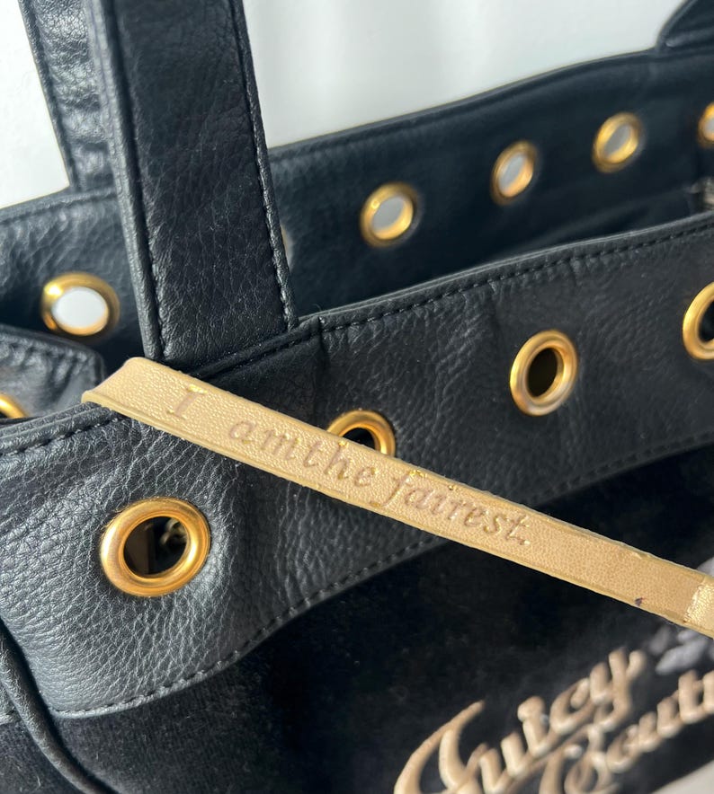 May include: Black tote bag with gold-toned grommets and handles. The bag features a gold leather tag with the inscription "I am the fairest." The brand name "Juicy Couture" is visible in gold lettering.