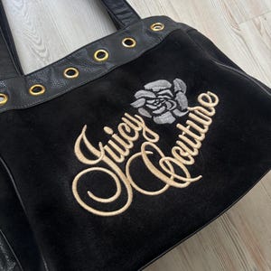 May include: Black tote bag with the embroidered text "Juicy Couture" and a rose graphic. The bag features black leather straps and gold-colored grommets along the top edge. The bag is made of a soft, velvety material.